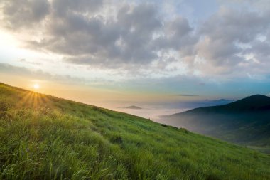 Dağ manzarası gündoğumu, güzel havalarda. Yeşil çimenli sarp bir tepe, sisli Vadisi ve ile parlak mavi gökyüzü altında uzak dağ Güneş beyaz bulutlar yükselterek yaktı. Doğa kavramının güzellik.