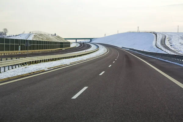 Modern wide smooth empty asphalt highway stretching to horizon under high bridge turning right past distant forest at day. Speed, comfortable journey and professional road building concept.