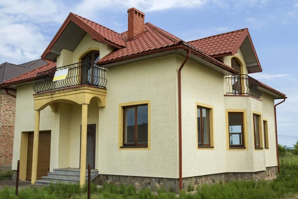 New yellow two-story residential cottage with shingle roof, front porch, plastic windows, high chimneys, balconies on blue sky background. Real estate property, architecture and prosperity concept.