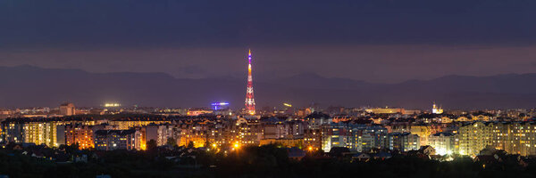 Wide panorama, aerial night view of modern tourist Ivano-Frankivsk city, Ukraine. Scene of bright lights of tall buildings, high television tower and green suburbs on Carpathian mountains background.