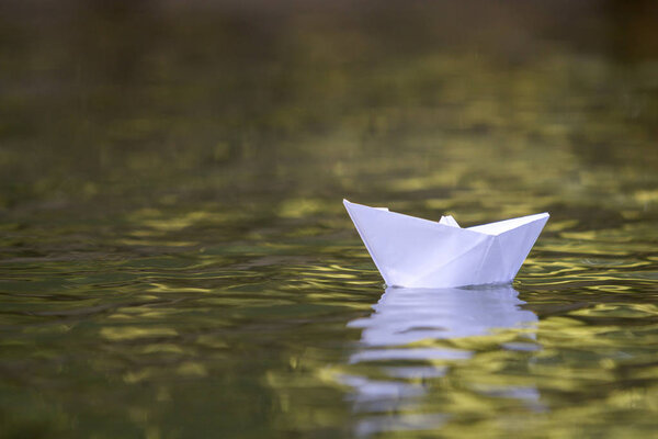 Close-up of simple small white origami paper boat floating quietly in yellow clear river or sea water under bright summer sky. Freedom, dreams and fantasies concept, copyspace background.