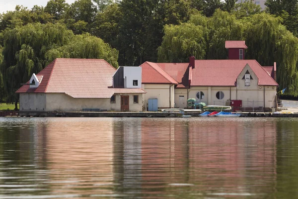 Water station building mooring on green willows background reflected in clear lake water surface on bright sunny summer day. Rental boats and catamarans for hire anchored at shore. Recreation concept.