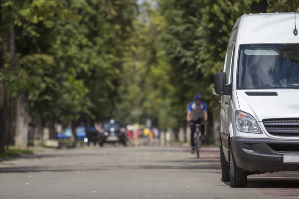 White passenger medium size commercial German luxury minibus van parked on city street with blurred silhouettes of pedestrians and moving car under green trees.