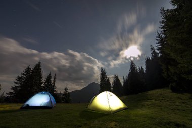 Parlak büyük ay koyu mavi bulutlu gökyüzü uzak dağ arka plan üzerinde uzun boylu çam ağaçları arasında takas yeşil çimenli ormanda iki turist çadır. Turizm, yaz dağda kamp gece.