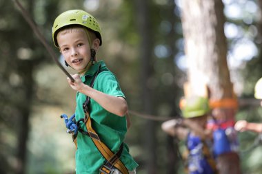 Genç Sevimli sarışın çocuk boy gülümseyerek emniyet kemeri ve karabina ile kablosuna bağlı kask ip yol boyunca parlak güneşli bokeh arka plan üzerinde taşır. Spor, oyun, eğlence, açık hava etkinliği konsepti.