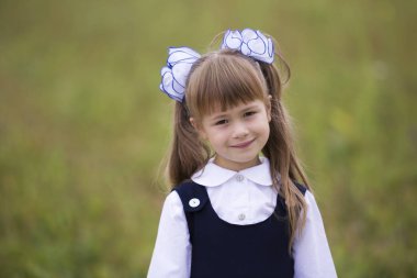 Yakın çekim portre sevimli tatlı gülümseyen ilk sınıf öğrencisi, küçük kız okul üniforması ve beyaz yay bulanık ışık yeşil kopya alan arka plan üzerinde uzun sarı saç. Eğitim ve okul kavramı.