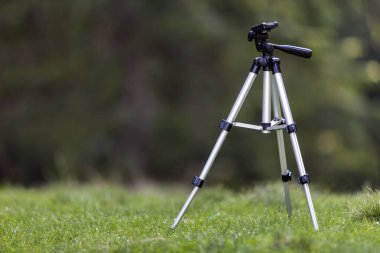 İzole alüminyum parlak tripod ayakta taze çimenli çayırda yalnız bulanık koyu yeşil orman ağaçları yeşillik bokeh kopya alan arka plan üzerinde parlak güneşli yaz gününde. İş ve hobi olarak fotoğraf.