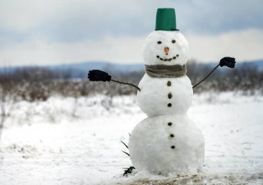 Büyük kova şapka, eşarp ve eldiven beyaz karlı alan kış manzara, bulanık siyah ağaçlar ve mavi gökyüzü kopya alan arka plan ile kardan adam gülümsüyor. . Mutlu Noeller ve mutlu yeni yıl tebrik kartı.