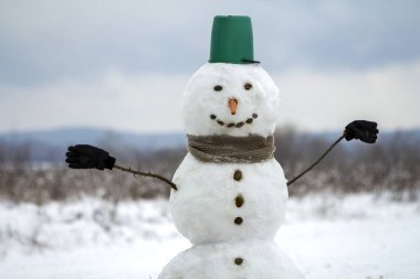 Büyük kova şapka, eşarp ve eldiven beyaz karlı alan kış manzara, bulanık siyah ağaçlar ve mavi gökyüzü kopya alan arka plan ile kardan adam gülümsüyor. . Mutlu Noeller ve mutlu yeni yıl tebrik kartı.