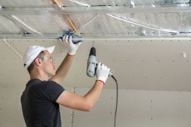Alçıpan sabitleme Gözlük Genç adam metal çerçeve parlak alüminyum folyo ile yalıtımlı tavanda Elektrikli tornavida kullanarak tavana askıya. Tadilat, inşaat, do It yourself konsepti.