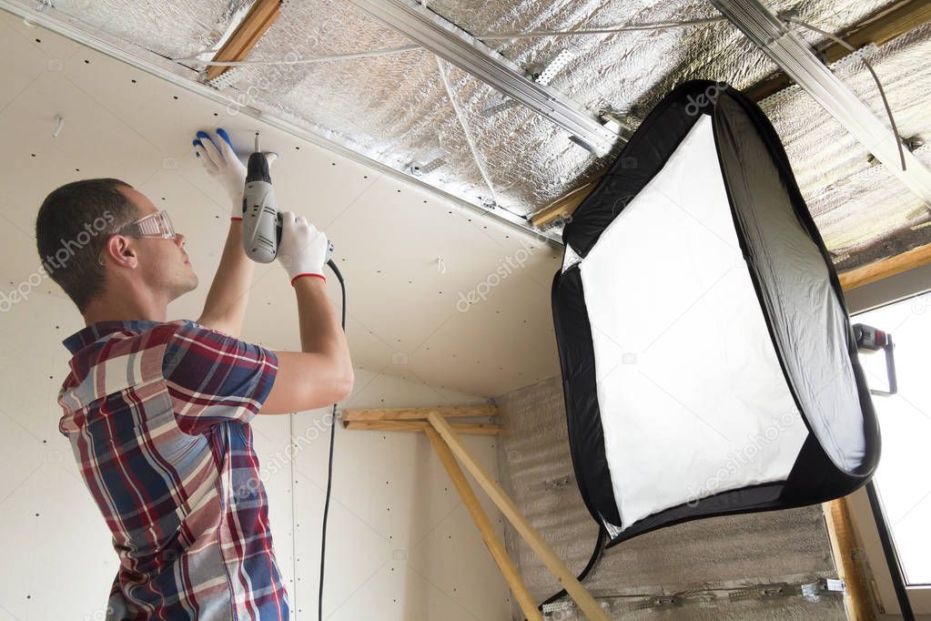 Young man in goggles and work gloves fixing drywall suspended ceiling to metal frame with electrical screwdriver on ceiling insulated with aluminum foil in front of soft box. Studio shot concept.