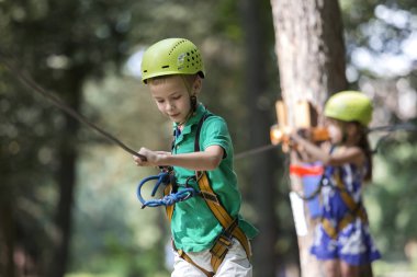 Genç Sevimli sarışın çocuk boy gülümseyerek emniyet kemeri ve karabina ile kablosuna bağlı kask ip yol boyunca parlak güneşli bokeh arka plan üzerinde taşır. Spor, oyun, eğlence, açık hava etkinliği konsepti.