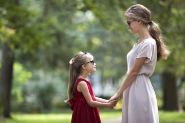 Profil genç sarışın uzun saçlı çekici gülümseyen kadın ve kız küçük çocuk güneş gözlüğü ve birbirlerine güneşli yaz yeşil bokeh özgeçmişlerine bakarak elele moda elbiseler.