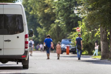 Beyaz yolcu orta boy ticari Lüks Minibüs Van arkadan görünüm n yaz şehir sokak üzerinde yeşil ağaç gölgeli bulanık siluetleri yayalar ve otomobil yeşil ağaçların altında park etmiş..