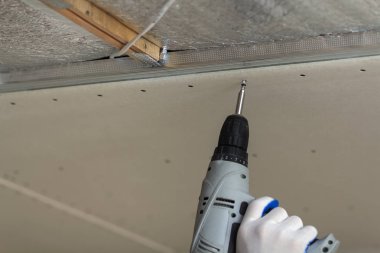 Oluşturucu işçi Close-up askıya alınmış Alçıpan Tavan metal çerçeveye bağlanma elektrik akülü tornavida ile koruyucu eldiven El. Yenileme, inşaat ve DIY kavramı.