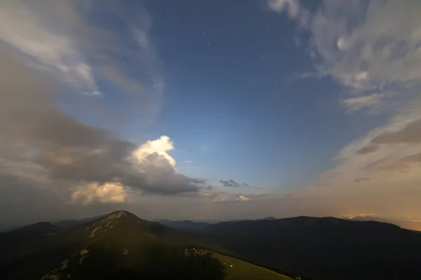 Beautiful summer night in mountains. Wide view of starry dark blue sky and lit by setting sun white clouds at sunset over magnificent woody mountain range, green grassy valley and dark pine trees.