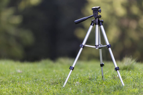 Isolated aluminum shiny tripod standing alone in fresh grassy meadow on bright sunny summer day on blurred dark green forest trees foliage bokeh copy space background. Photography as job and hobby.