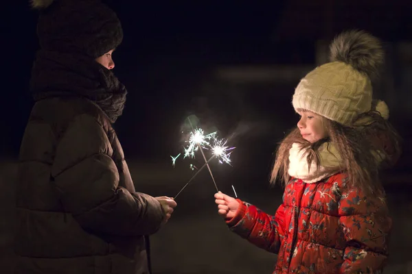 轮廓两个可爱的小孩 男孩和女孩在温暖的冬衣举行燃烧的火花烟花在黑暗的夜晚户外复制空间背景 新年和圣诞节庆祝概念 — 图库照片