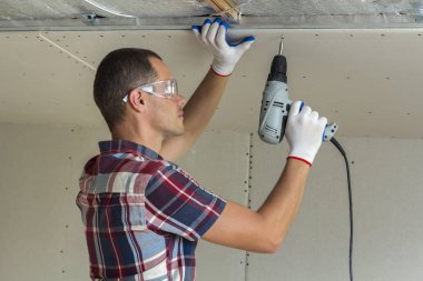 Alçıpan sabitleme Gözlük Genç adam metal çerçeve parlak alüminyum folyo ile yalıtımlı tavanda Elektrikli tornavida kullanarak tavana askıya. Tadilat, inşaat, do It yourself konsepti.