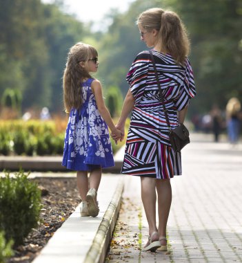 Genç sarışın uzun saçlı ince çekici kadın ve kız küçük çocuk güneş gözlüğü ve şık elbiseler güneşli yaz park uğrayacağız tutuşup yürürken görünümünü geri. Mutlu aile ilişkileri kavramı.