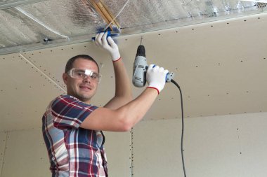Alçıpan sabitleme Gözlük Genç adam metal çerçeve parlak alüminyum folyo ile yalıtımlı tavanda Elektrikli tornavida kullanarak tavana askıya. Tadilat, inşaat, do It yourself konsepti.