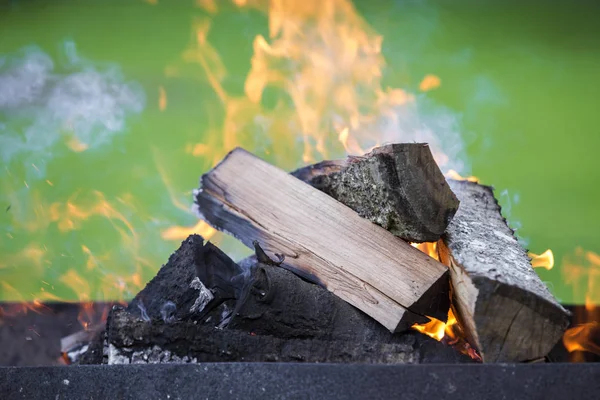 Brightly burning in metal box logs firewood for barbecue outdoor on sunny day. Orange high flame and white smoke on blurred blue sky and green grass background. Camping, safety and tourism concept.