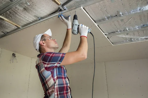 Young man in usual clothing and work gloves fixing drywall suspended ceiling to metal frame using electrical screwdriver on ceiling insulated with shiny aluminum foil. DIY, do it yourself concept.
