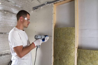 Gözlük yalıtımı çalışma tornavida ile işçi. Alçıpan Duvar kirişler, taş yünü personel ahşap çerçeve içinde yalıtım üzerinde. Rahat sıcak ev, ekonomi, inşaat ve tadilat kavramı.