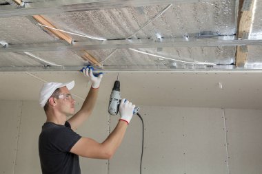 Alçıpan sabitleme Gözlük Genç adam metal çerçeve parlak alüminyum folyo ile yalıtımlı tavanda Elektrikli tornavida kullanarak tavana askıya. Tadilat, inşaat, do It yourself konsepti.