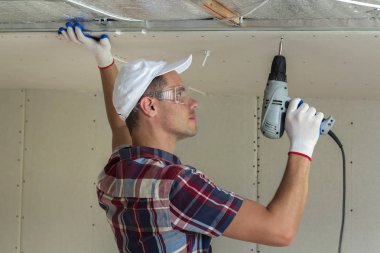 Alçıpan sabitleme Gözlük Genç adam metal çerçeve parlak alüminyum folyo ile yalıtımlı tavanda Elektrikli tornavida kullanarak tavana askıya. Tadilat, inşaat, do It yourself konsepti.
