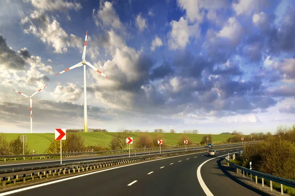 Wide highway with moving car stretching to horizon by green hills with high wind turbines on blue cloudy sky background. Modern technologies, renewable energy source and ecological problems solving.