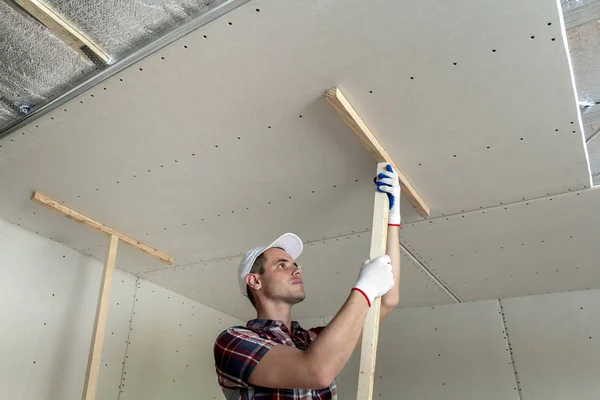 年轻工人在保护工作手套固定木制持有者的墙上悬挂天花板 金属框绝缘的光泽铝箔 Diy 做自己的概念 — 图库照片