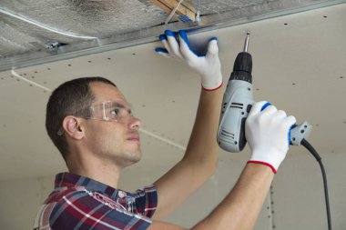 Alçıpan sabitleme Gözlük Genç adam metal çerçeve parlak alüminyum folyo ile yalıtımlı tavanda Elektrikli tornavida kullanarak tavana askıya. Tadilat, inşaat, do It yourself konsepti.