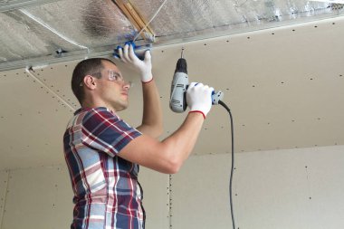 Alçıpan sabitleme Gözlük Genç adam metal çerçeve parlak alüminyum folyo ile yalıtımlı tavanda Elektrikli tornavida kullanarak tavana askıya. Tadilat, inşaat, do It yourself konsepti.