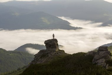 Atletik dağcı turist dağ Vadisi üzerinde yüksek kayalık oluşumu üzerinde silueti beyaz bulutlar ve sis ile dolu ve herdem yeşil orman dağ yamaçları altında açık gökyüzü arka plan ile kaplı.