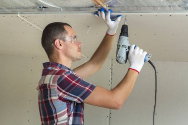 Alçıpan sabitleme Gözlük Genç adam metal çerçeve parlak alüminyum folyo ile yalıtımlı tavanda Elektrikli tornavida kullanarak tavana askıya. Tadilat, inşaat, do It yourself konsepti.