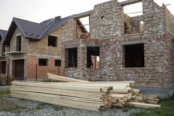 Future cottage under construction and pile of gravel and stack of boards in front of not finished new big brick house with brown shingled roof. Construction, investment and real property concept.