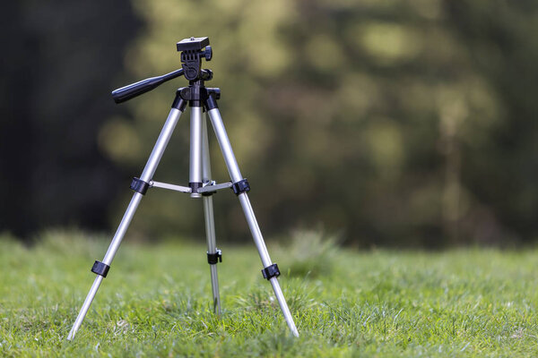 Isolated aluminum shiny tripod standing alone in fresh grassy meadow on bright sunny summer day on blurred dark green forest trees foliage bokeh copy space background. Photography as job and hobby.
