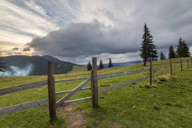 Yeşil çimenli Vadisi yamaç ilkel ahşap çit ve kapı ile panoramik yaz görünümünü gündoğumu, mavi bulutlu gökyüzü altında uzak odunsu sisli dağda. Güzellik, Doğa ve kavram seyahat.