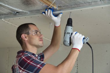 Alçıpan sabitleme Gözlük Genç adam metal çerçeve parlak alüminyum folyo ile yalıtımlı tavanda Elektrikli tornavida kullanarak tavana askıya. Tadilat, inşaat, do It yourself konsepti.