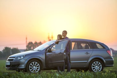 Genç çift, çekici ince uzun - saçlı kadın ve yakışıklı adam birlikte gündoğumu veya günbatımı parlak gökyüzü üzerinde kırsal kırsalında açık kapı ile gümüş renkli araba, ayakta kopyalama alanı arka plan.