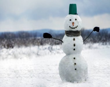 Büyük kova şapka, eşarp ve eldiven beyaz karlı alan kış manzara, bulanık siyah ağaçlar ve mavi gökyüzü kopya alan arka plan ile kardan adam gülümsüyor. . Mutlu Noeller ve mutlu yeni yıl tebrik kartı.