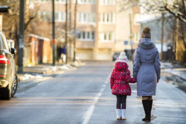 Arkadan görünüşü genç ince çekici kadın anne ve küçük çocuk kız kızı sıcak giysiler birlikte şehir boyunca güneşli kış gününde sokak üzerinde kentsel arka plan bulanık elele yürüyüş.