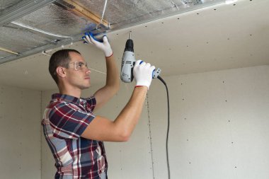 Alçıpan sabitleme Gözlük Genç adam metal çerçeve parlak alüminyum folyo ile yalıtımlı tavanda Elektrikli tornavida kullanarak tavana askıya. Tadilat, inşaat, do It yourself konsepti.