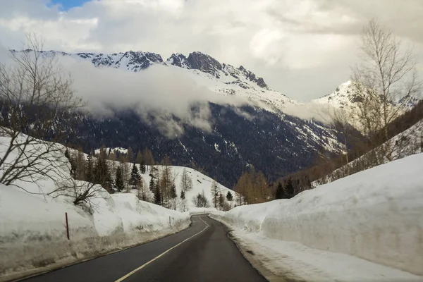 Empty twisty asphalt road stretching through deep snow towards beautiful bright white Alps steep rocky woody snowy mountain peaks under cloudy winter sky. Winter landscape on cold sunny day.