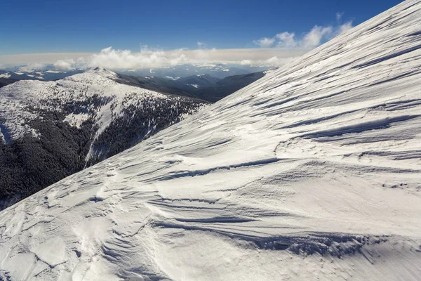 Beautiful winter landscape. Steep mountain hill slope with white deep snow, distant woody mountain range panorama stretching to horizon and bright shining sun rays on blue sky copy space background.