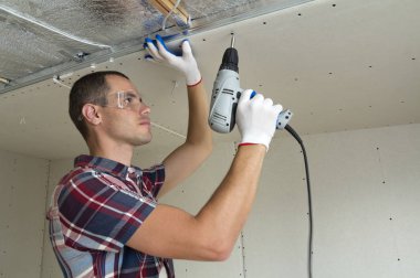 Alçıpan sabitleme Gözlük Genç adam metal çerçeve parlak alüminyum folyo ile yalıtımlı tavanda Elektrikli tornavida kullanarak tavana askıya. Tadilat, inşaat, do It yourself konsepti.
