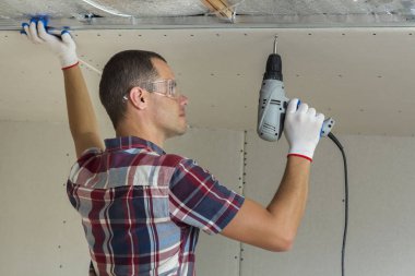 Alçıpan sabitleme Gözlük Genç adam metal çerçeve parlak alüminyum folyo ile yalıtımlı tavanda Elektrikli tornavida kullanarak tavana askıya. Tadilat, inşaat, do It yourself konsepti.