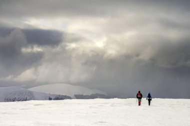 Winter mountain landscape. Back view of travelers tourist hikers with backpacks on snowy field walking towards distant mountain on cloudy dark blue stormy sky copy space background on cold winter day. clipart