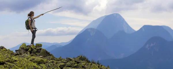 Young slim blond tourist girl with backpack points with stick at foggy mountain range panorama standing on rocky top on bright blue morning sky background. Tourism, traveling and climbing concept.